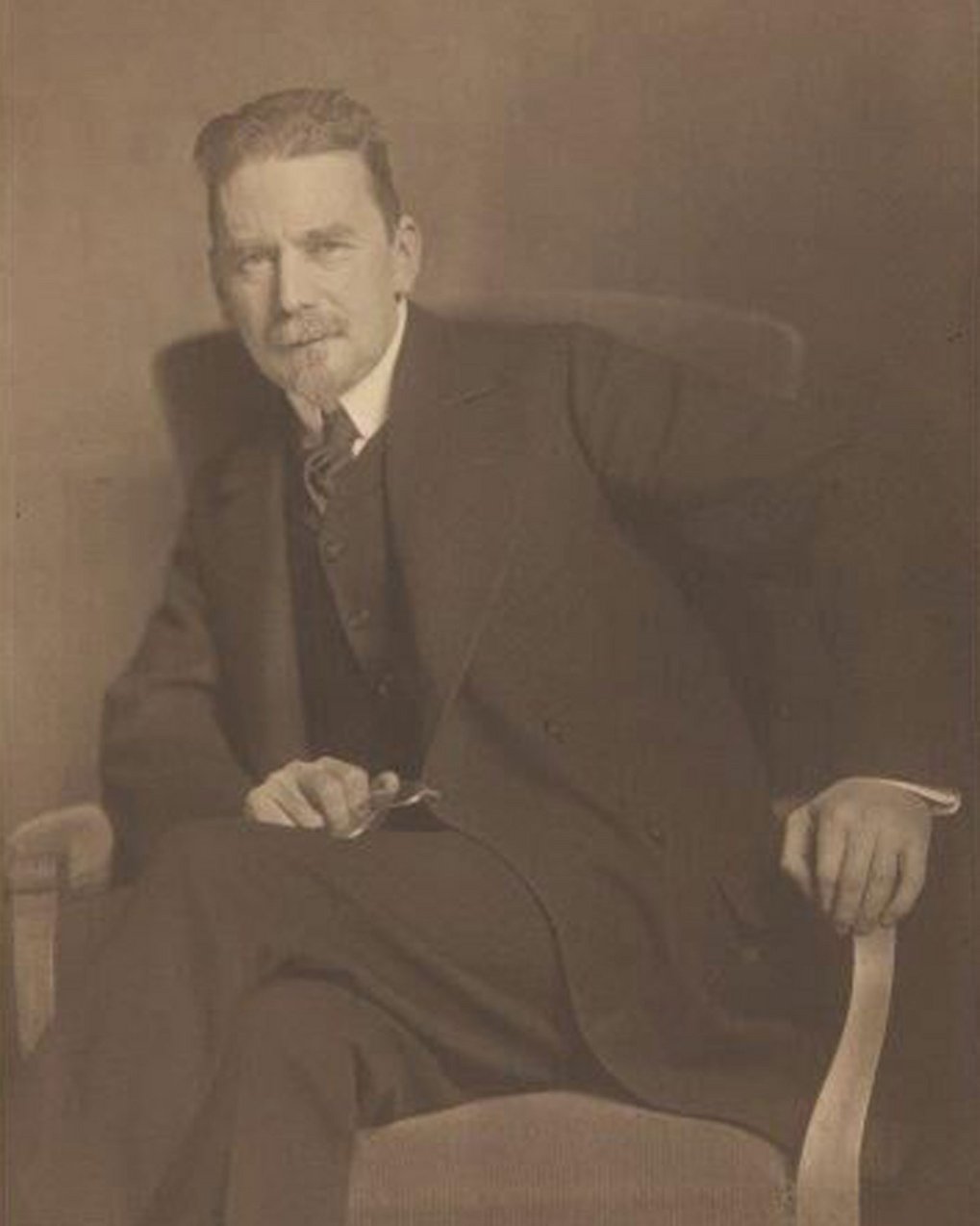 Man in formal suit sitting on a wooden chair, holding a pair of glasses.