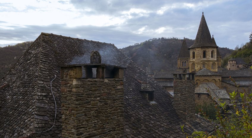 Conques in the Global World (abgeschlossen) Historische Steindächer und Schornsteine, Turm ragt hinter Hügeln hervor.
