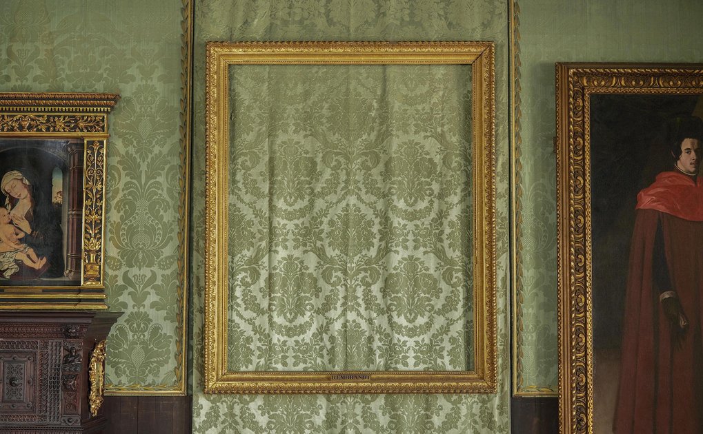 An empty gold frame hangs against green patterned wallpaper, flanked by two paintings.