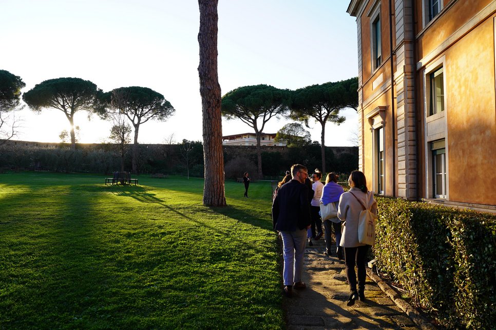 People walking near a building in a sunlit park, surrounded by trees.