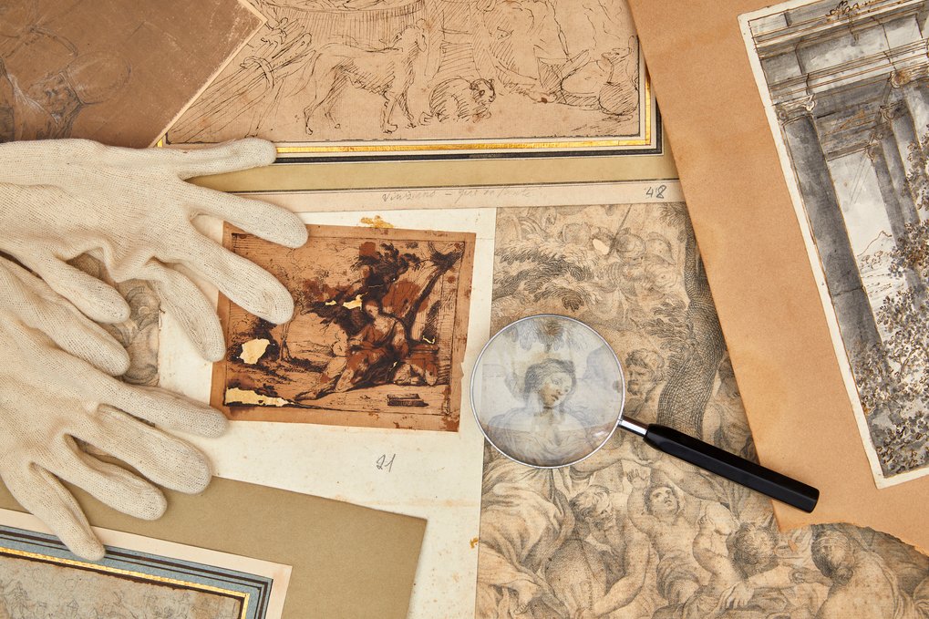 Diverse historische Zeichnungen, eine Lupe und weiße Handschuhe liegen auf einem Tisch.